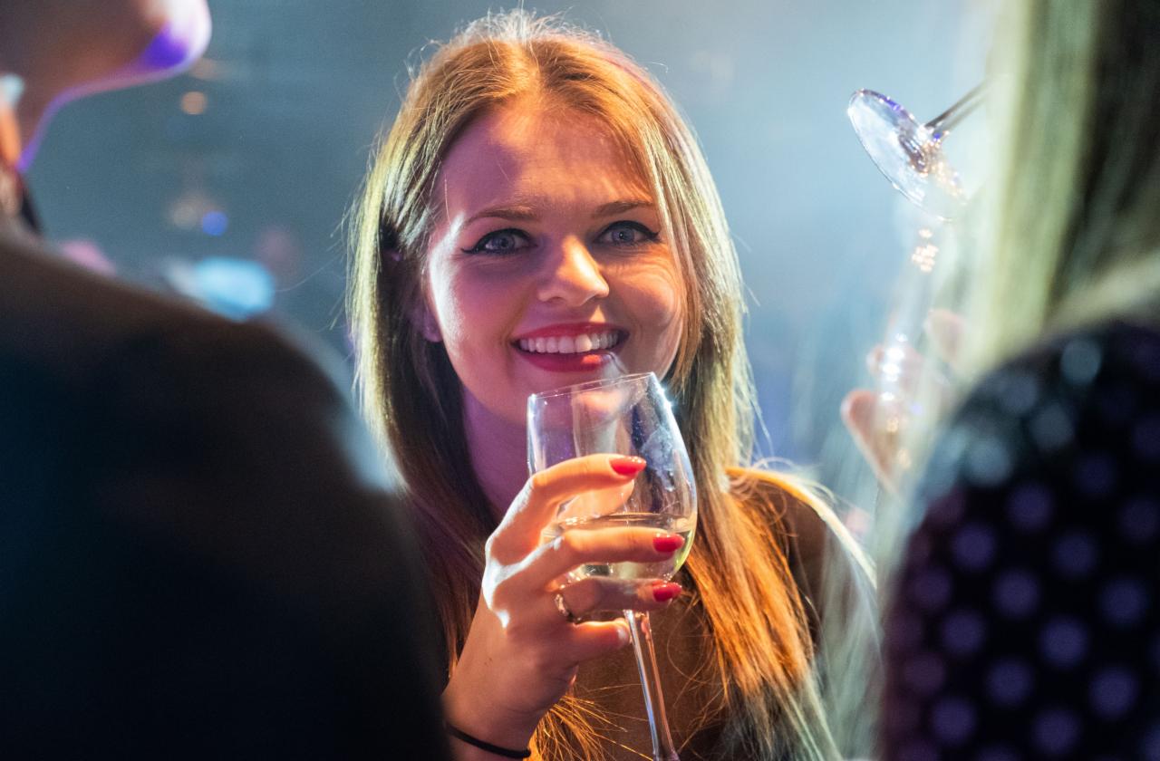Uśmiechnięta kobieta z długimi włosami, trzymająca kieliszek białego wina w dłoni z czerwonymi paznokciami, w tle rozmyte postacie i oświetlenie imprezowe.