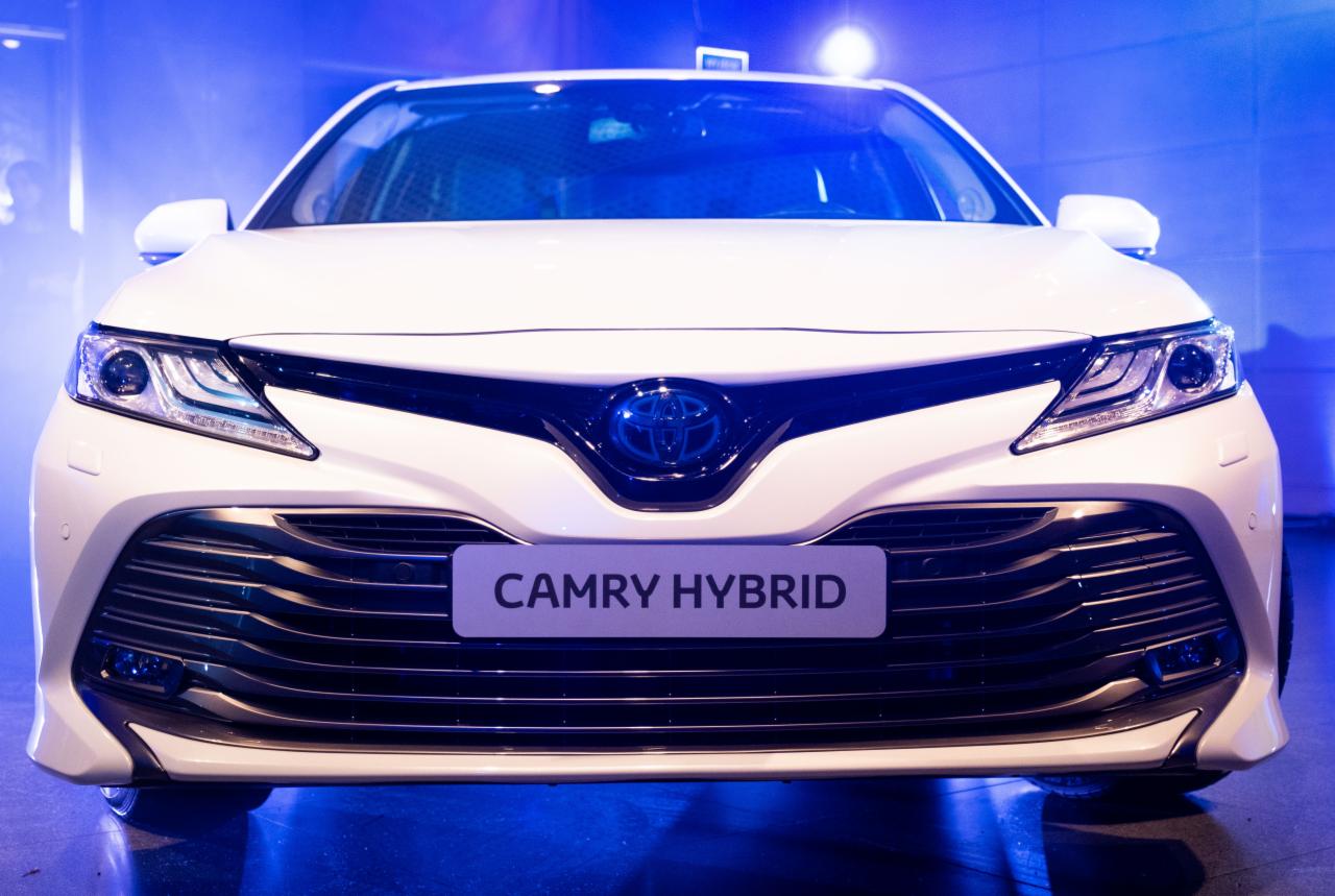 Przód białego samochodu Toyota Camry Hybrid z wyraźnie widocznym logo na tle niebieskiego oświetlenia.