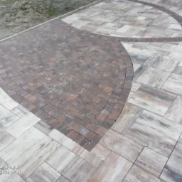 Ułożona nawierzchnia z kostki brukowej w dwóch odcieniach brązu i beżu, z dekoracyjnym wzorem w kształcie łuku i półkola, widoczne nierówności terenu w tle.