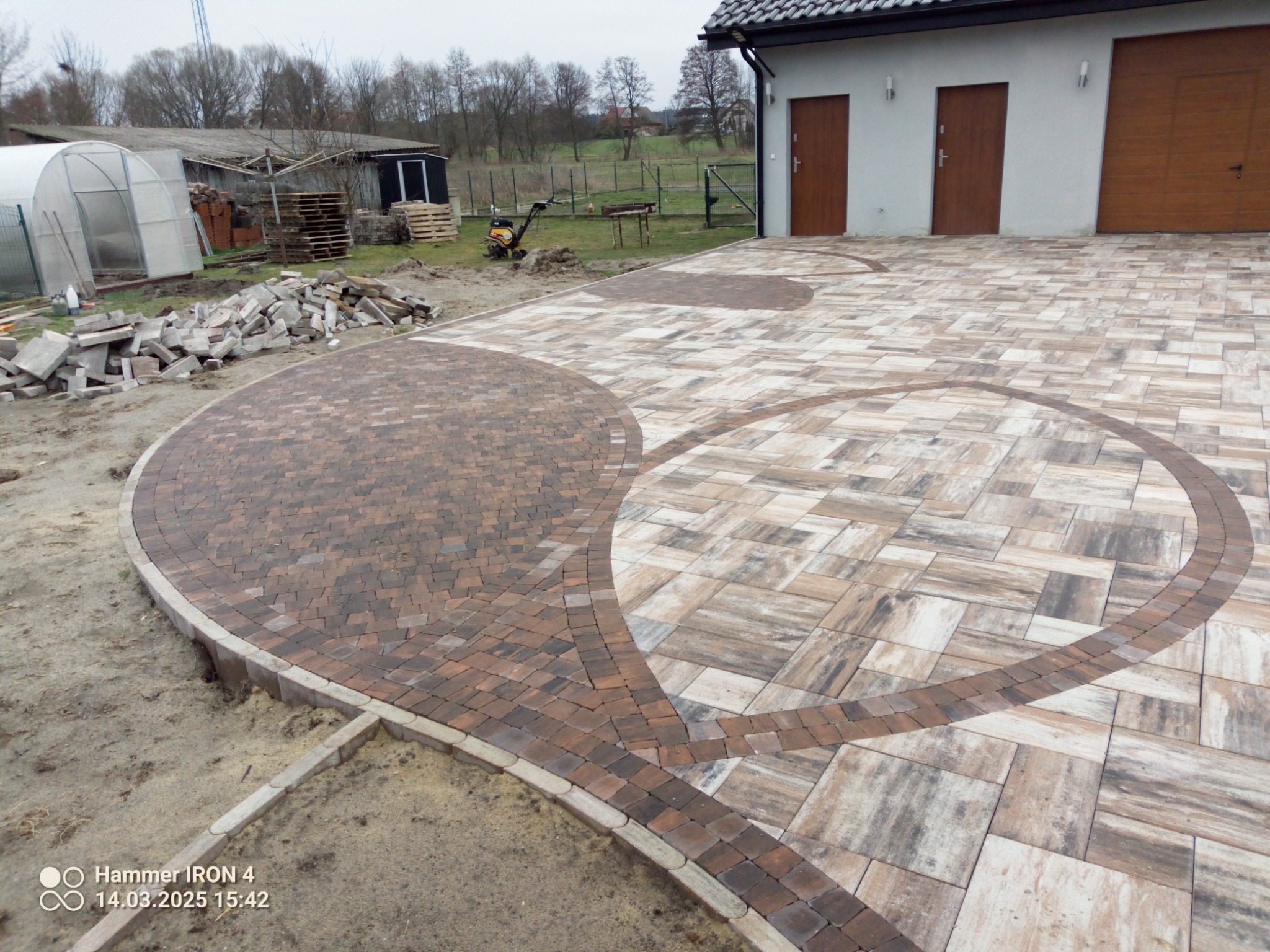 Ułożona kostka brukowa w kształcie dwóch łez, z obrzeżami z ciemniejszej kostki, na tle budynku z dwiema parami drzwi i bramą garażową, widoczny stos kostki do dalszych prac.