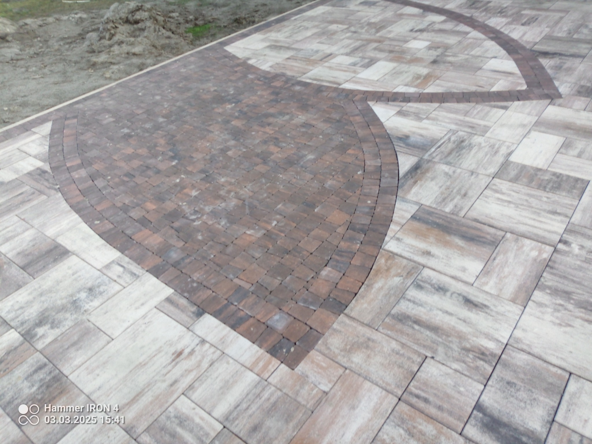 Ułożona nawierzchnia z kostki brukowej w dwóch odcieniach brązu i beżu, z dekoracyjnym wzorem w kształcie łuku i półkola, widoczne nierówności terenu w tle.