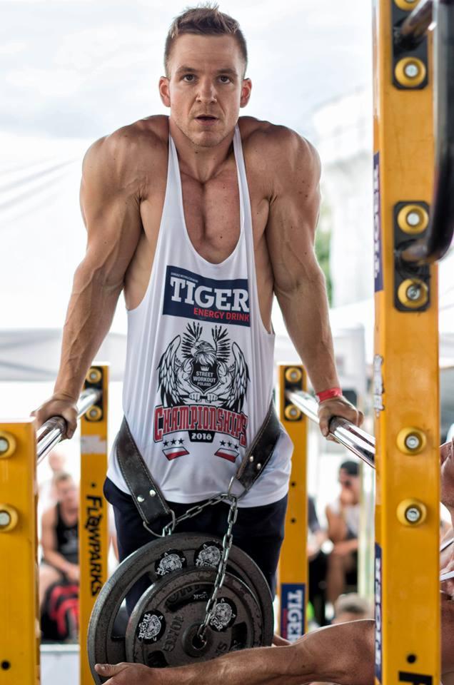 Mężczyzna w koszulce z logo Tiger Energy Drink, wykonujący ćwiczenie na poręczach z obciążeniem, skupiony wzrok, widoczne żyły na ramionach.