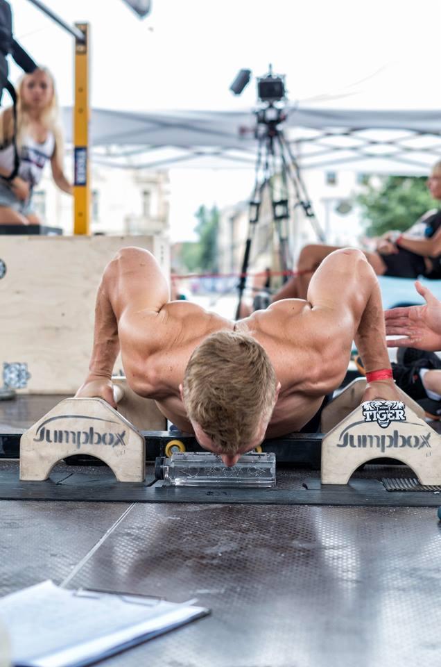 Mężczyzna wykonuje pompki na podwyższeniach Jumpbox, eksponując muskulaturę ramion na tle sceny z widocznym sprzętem nagłośnieniowym i publicznością.