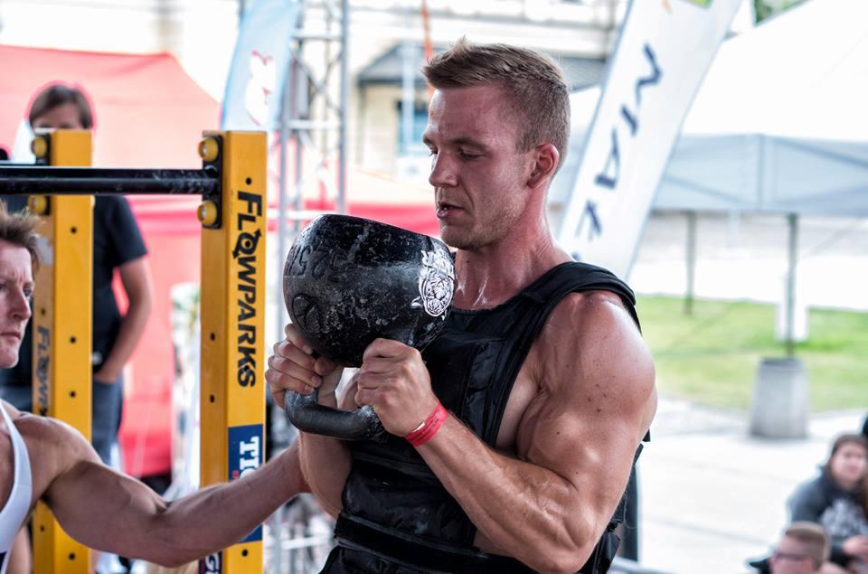 Mężczyzna w kamizelce obciążeniowej podnosi ciężki kettlebell na tle żółtego słupka z napisem Flowparks, ćwiczenia na świeżym powietrzu.