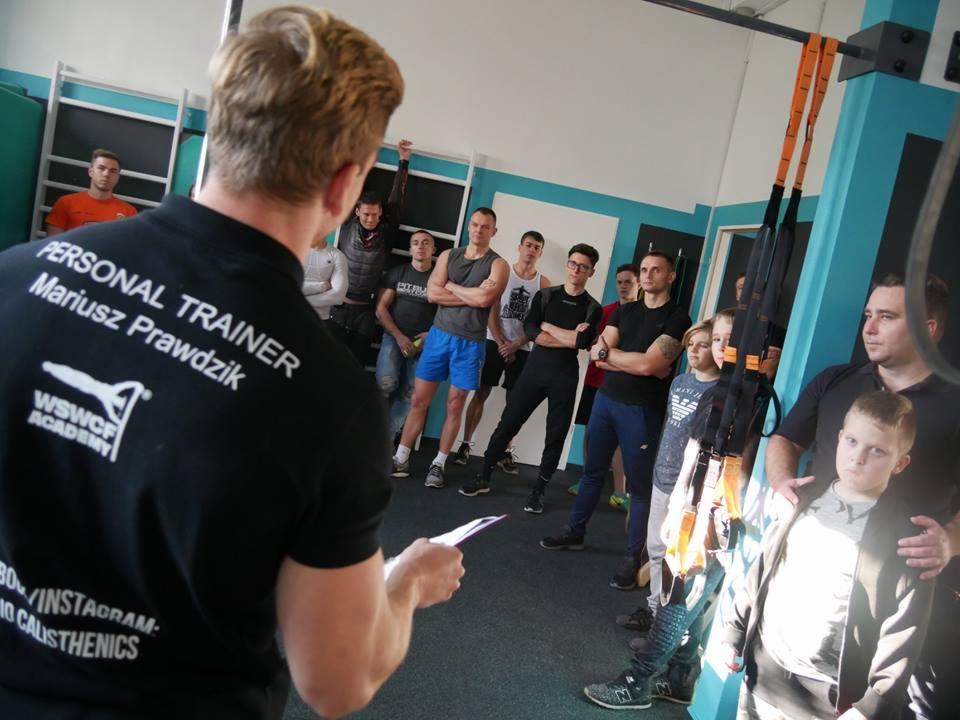 Instruktor w czarnej koszulce z napisem 'PERSONAL TRAINER Mariusz Prawdzik WSWCF ACADEMY' trzymający kartkę, przemawia do grupy osób w sali z przyrządami do ćwiczeń.