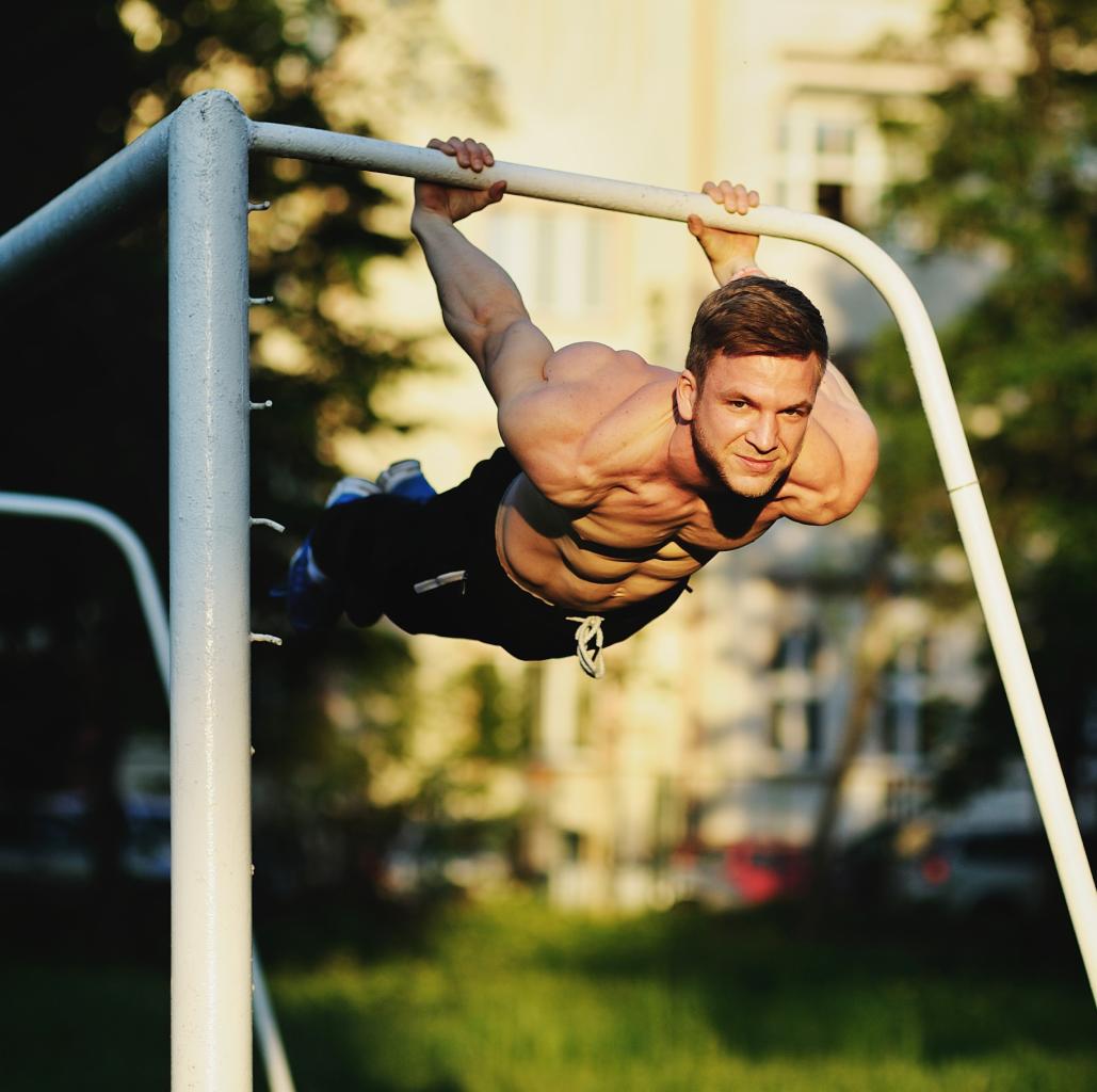 Mężczyzna wykonujący ćwiczenie siłowe na drążku na świeżym powietrzu, z budynkiem w tle.