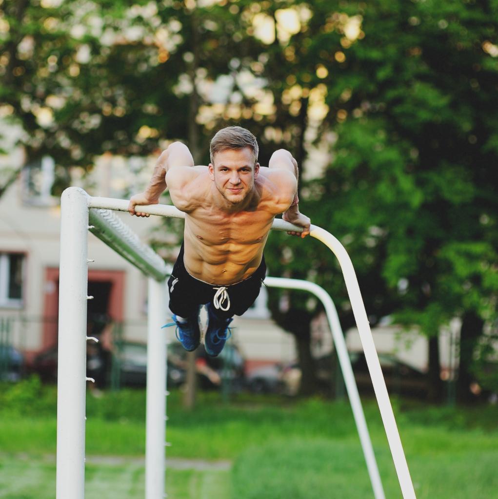 Mężczyzna bez koszulki wykonuje ćwiczenie siłowe na drążku na zewnątrz, widoczne mięśnie brzucha i ramion, w tle budynek i zieleń.