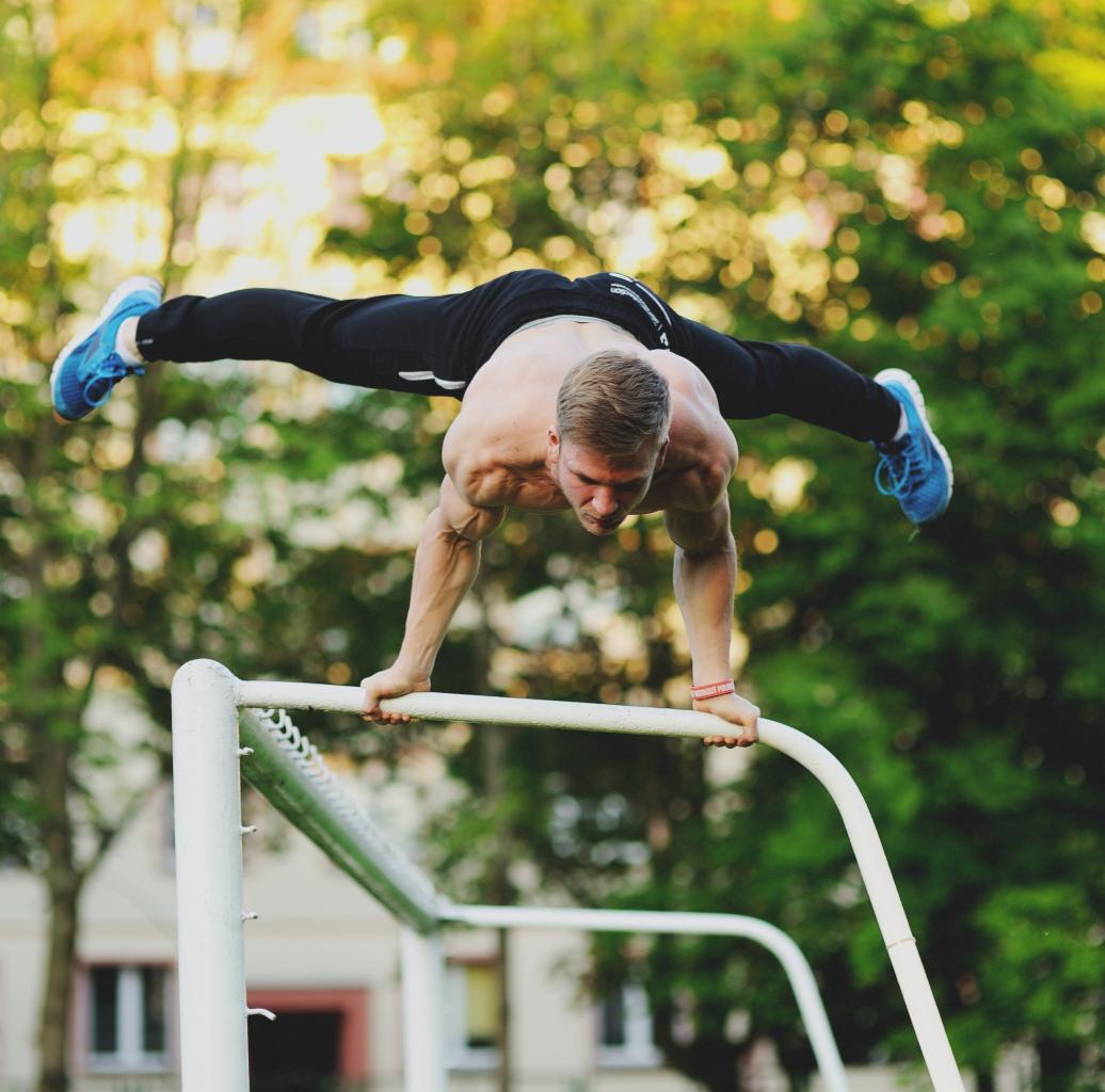 Mężczyzna bez koszulki w czarnych spodniach i niebieskich butach wykonuje szpagat w staniu na rękach na białej bramce do piłki nożnej na tle zieleni.