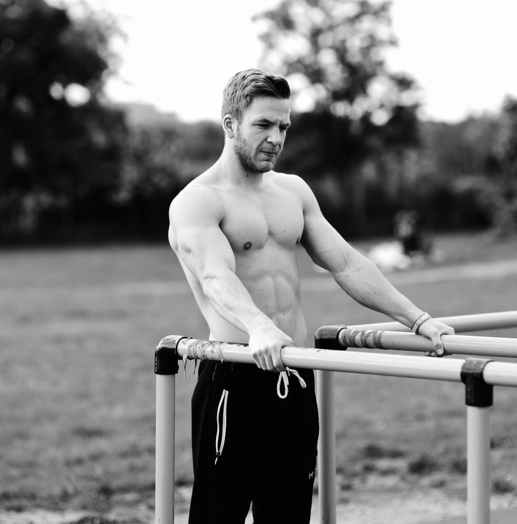 Mężczyzna bez koszulki, z widocznymi mięśniami brzucha, opiera się o metalowe poręcze na tle parku, zdjęcie czarno-białe.
