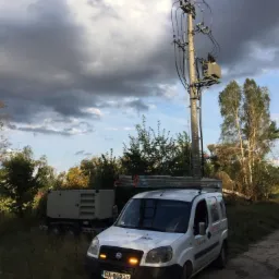 Biały samochód dostawczy z drabiną na dachu zaparkowany obok słupa energetycznego z transformatorem, w tle agregat prądotwórczy i zachmurzone niebo.