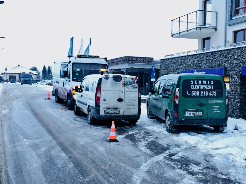 Flota pojazdów serwisowych zaparkowana na ośnieżonej ulicy; biała furgonetka z napisem 'Instalacje Elektryczne Monitoringowe' i zielony van z napisem 'Serwis Elektryczny' widoczne z tyłu, ustawione...
