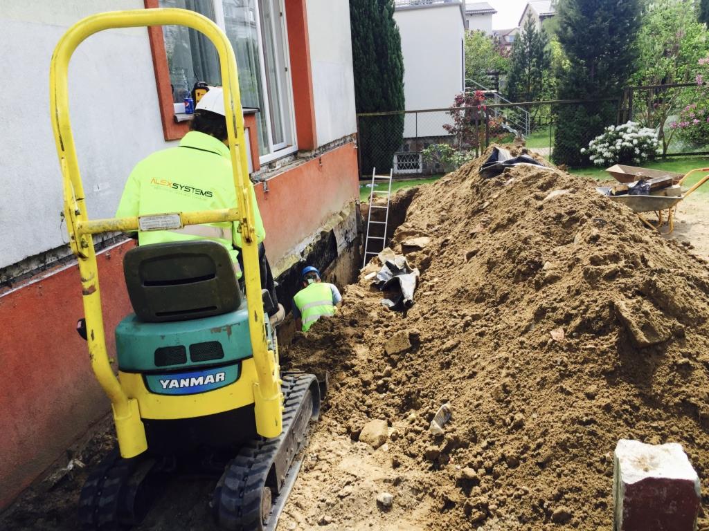 Prace ziemne przy fundamentach budynku, widoczny mini-koparka Yanmar, wykopany rów i sterta ziemi, dwóch pracowników w kaskach i kamizelkach odblaskowych pracuje w wykopie, drabina oparta o ścianę...