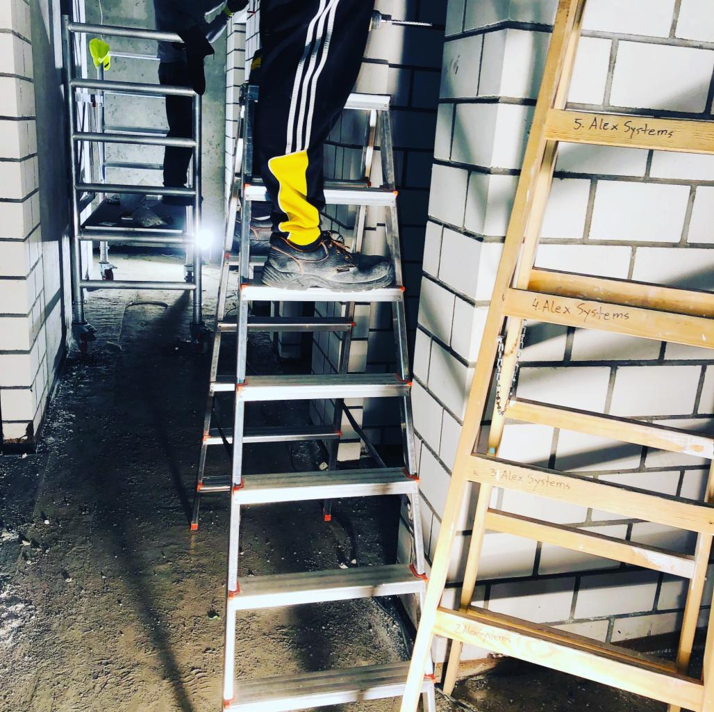 Pracownik w czarno-żółtych spodniach na aluminiowej drabinie w surowym wnętrzu, obok drewniana drabina z napisem 'Alex Systems', w tle rusztowanie.