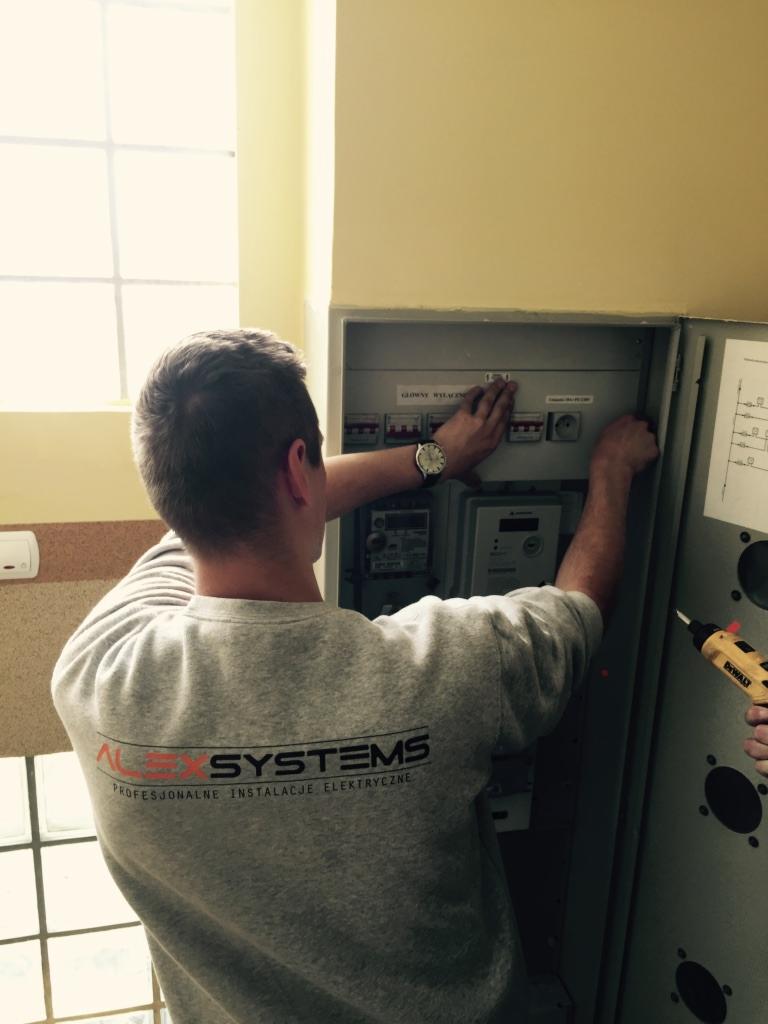 Elektryk w szarej bluzie z logo 'Alex Systems' sprawdza instalację elektryczną w szafce rozdzielczej, widoczny zegarek na ręce i wkrętarka DeWalt.