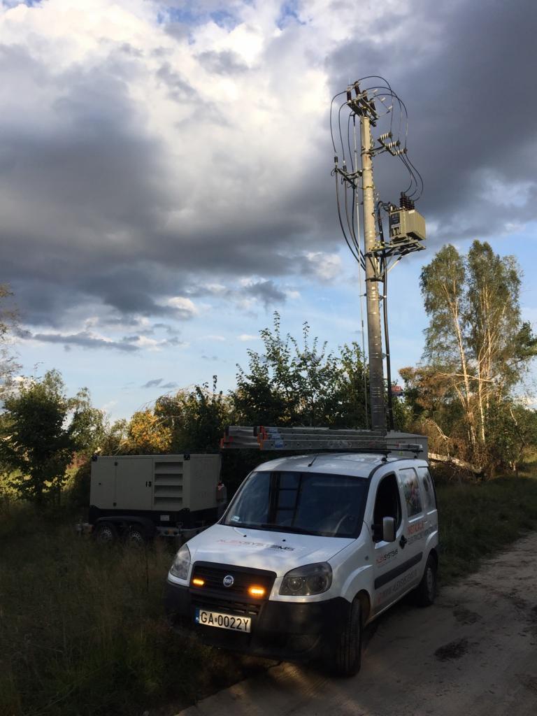 Biały samochód dostawczy z drabiną na dachu zaparkowany obok słupa energetycznego z transformatorem, w tle agregat prądotwórczy i zachmurzone niebo.