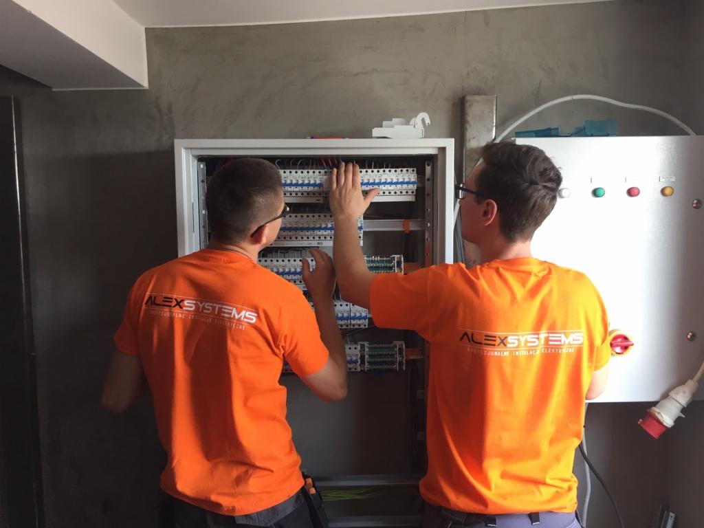 Dwóch mężczyzn w pomarańczowych koszulkach z logo ALEXSYSTEMS pracuje przy rozdzielnicy elektrycznej w szarej, surowej przestrzeni.