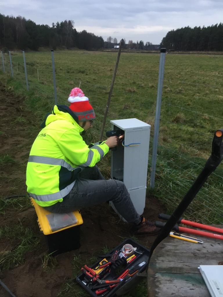 Pracownik w jaskrawej kurtce i czapce z pomponem, siedząc na żółtym pudełku, instaluje kable w szarej skrzynce elektrycznej na zewnątrz, z widocznymi narzędziami i polami w tle.