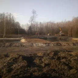 Krajobraz po pracach ziemnych: wykopane stawy otoczone lasem, widoczny ciężki sprzęt - koparka i spychacz, ziemia nierówna, bez roślinności, w tle drzewa bez liści pod jasnym niebem.