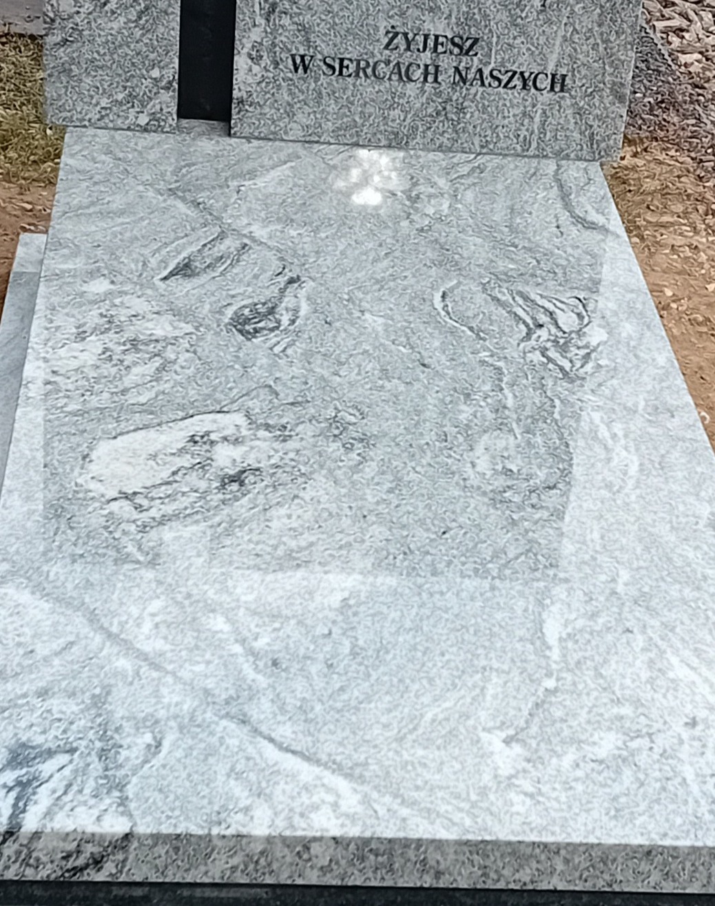 Jasnoszary nagrobek z marmurową płytą, na której widnieje napis 'Żyjesz w sercach naszych', leżący na ziemi.