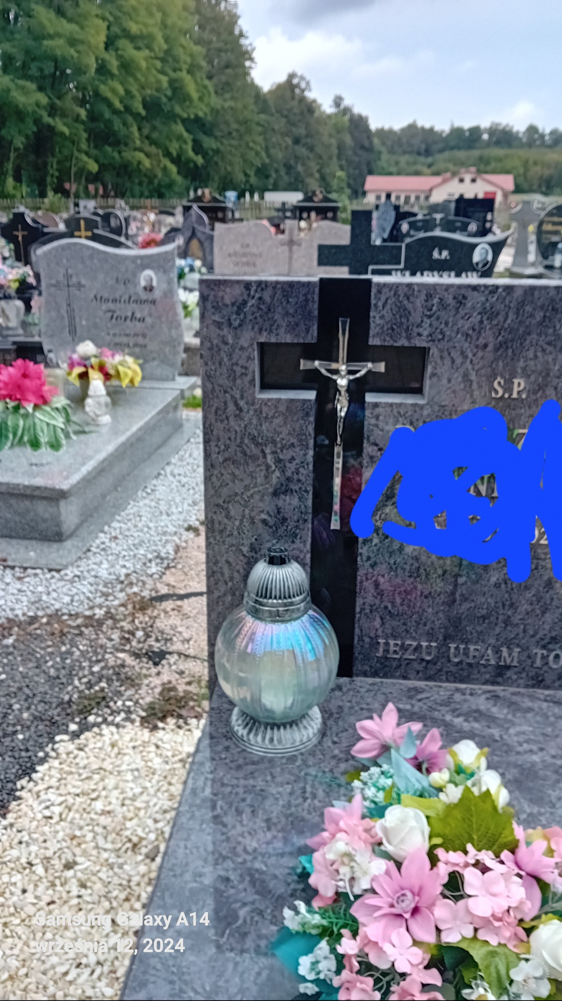 Granitowy nagrobek z krzyżem i figurą Jezusa, lampionem i kwiatami na cmentarzu, z widocznymi innymi nagrobkami w tle.