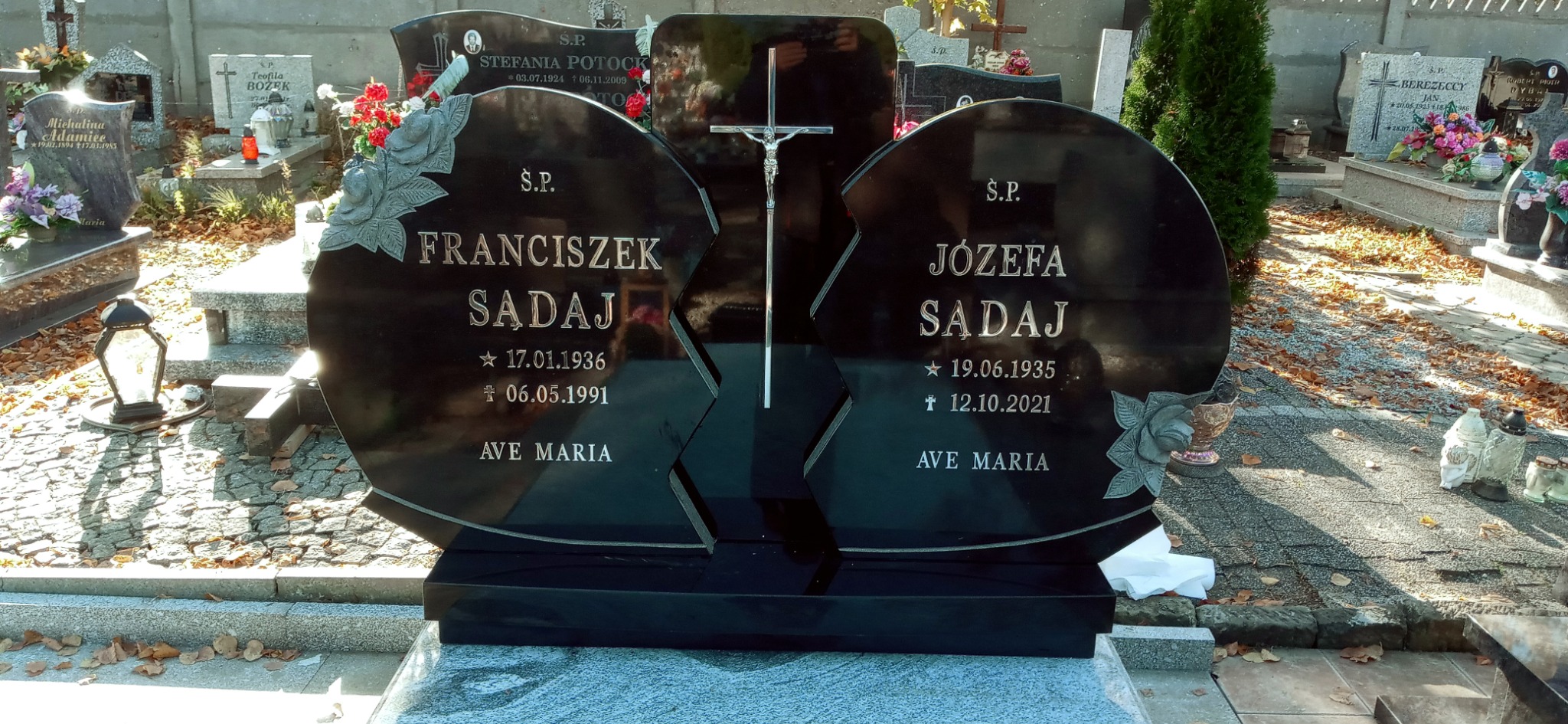 Czarny, podwójny nagrobek z wygrawerowanymi różami, imionami Franciszek i Józefa Sadaj oraz datami urodzin i śmierci, na tle cmentarza z innymi nagrobkami i opadłymi liśćmi.