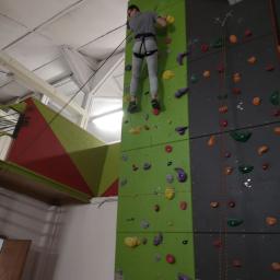 Mobilna Myjnia Mycie Pełną Parą - Mężczyzna wspina się na ściankę wspinaczkową z kolorowymi uchwytami, zabezpieczony liną. Widoczny fragment sufitu hali i sąsiednia ściana.