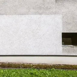 Fragment ściany budynku z wyraźnie odróżniającym się, wyczyszczonym prostokątnym obszarem, sąsiadującym z brudną powierzchnią i małym, ciemnym otworem okiennym, z zielonym trawnikiem u podstawy.