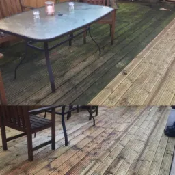 Porównanie czyszczenia drewnianego tarasu: połowa pokryta zielonym nalotem, druga połowa świeżo umyta, ukazująca naturalny kolor drewna, z widocznym stołem i krzesłami na tarasie.