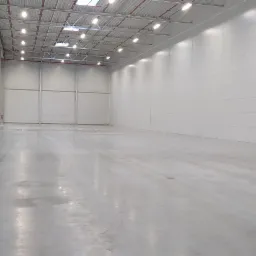 Wnętrze hali magazynowej z betonową podłogą i białymi ścianami panelowymi, oświetlone lampami sufitowymi, z pracownikiem w pomarańczowej kamizelce w oddali i czarnym odkurzaczem przemysłowym przy...