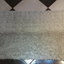 Beżowy, puszysty dywanik shaggy leżący na podłodze w szachownicę z płytek ceramicznych w odcieniach beżu i brązu.