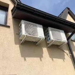 Dwa białe klimatyzatory zewnętrzne marki Vesser zamontowane na beżowej elewacji domu pod brązowym dachem i rynną.