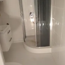 Wnętrze łazienki w trakcie remontu: widoczna kabina prysznicowa z brudną podłogą, umywalka z szafką i fragment toalety. Białe płytki na ścianach i podłodze. Widoczne ślady po pracach remontowych.