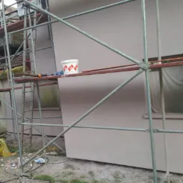 Elewacja budynku w trakcie malowania, widoczne rusztowanie, wiadro z farbą na podeście, kabel elektryczny na ziemi.