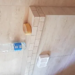 Beżowe płytki trawertyn na podłodze i ścianie w łazience, fuga z małych płytek, gąbka, butelka wody i biała kostka. Widoczne elementy instalacji.