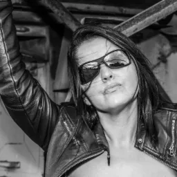 Czarno-białe zdjęcie kobiety w skórzanej kurtce i okularach przeciwsłonecznych, z wiatrem we włosach, pozującej na tle metalowej konstrukcji.