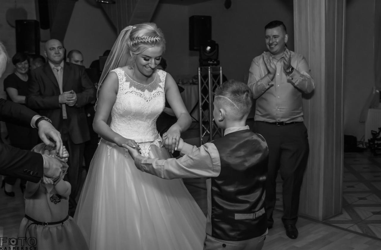 Czarno-białe zdjęcie panny młodej w koronkowej sukni, tańczącej z chłopcem podczas wesela, z uśmiechniętymi gośćmi w tle.