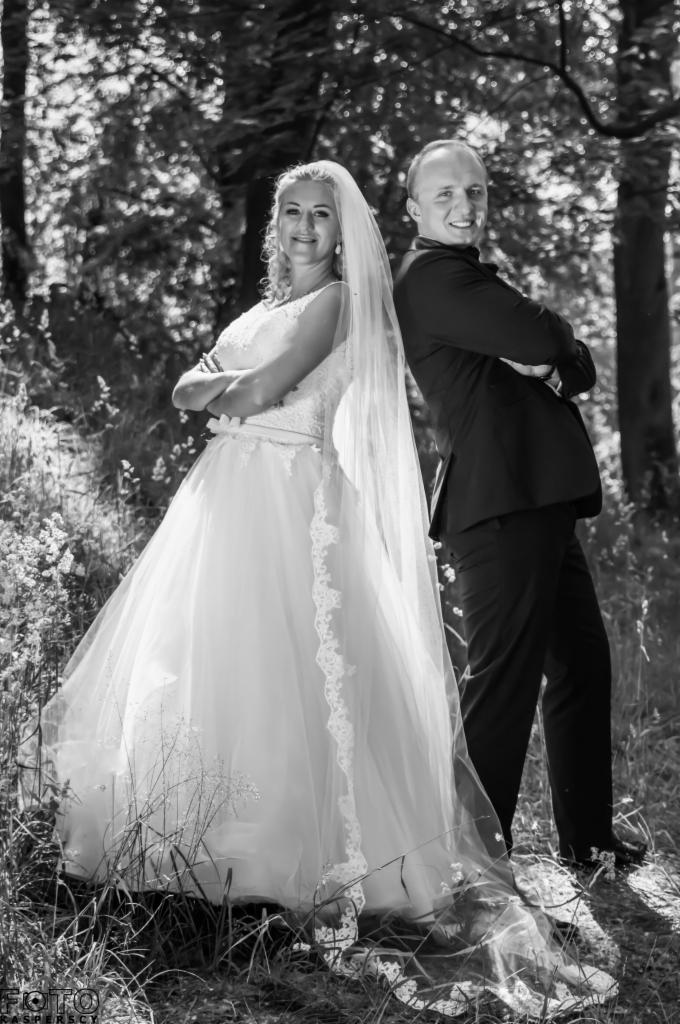 Czarno-białe zdjęcie pary młodej stojącej plecami do siebie w lesie, panna młoda w koronkowej sukni i długim welonie, pan młody w czarnym garniturze, oboje z uśmiechem i skrzyżowanymi ramionami.