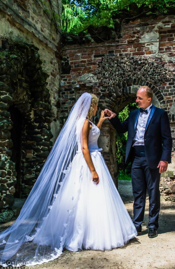 Para młoda w ruinach zamku: Panna młoda w białej sukni i welonie, pan młody w granatowym garniturze, dłonie ułożone w kształt serca na tle ceglanej ściany i zieleni.