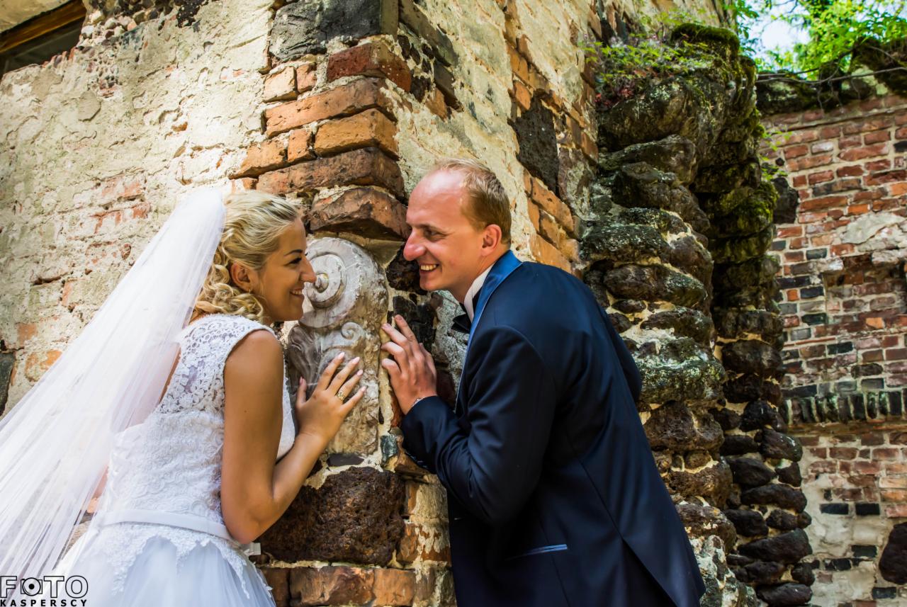 Uśmiechnięta para młoda wpatrzona w siebie, opierająca się o starą ceglaną ścianę z dekoracyjnym elementem architektonicznym i roślinnością.