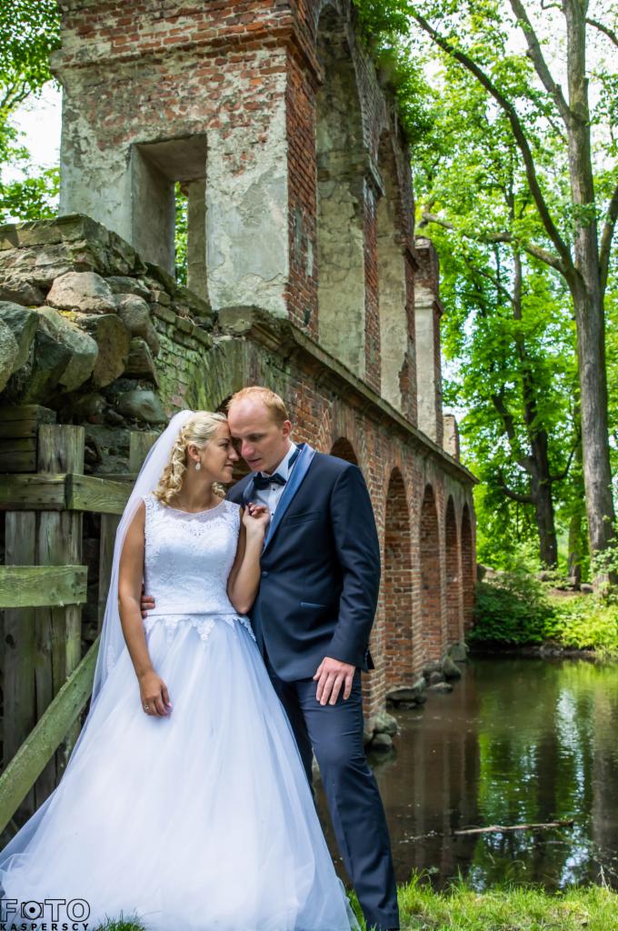 Para młoda w objęciach na tle zrujnowanej, ceglanej budowli z arkadami i stawem, otoczona zielenią.