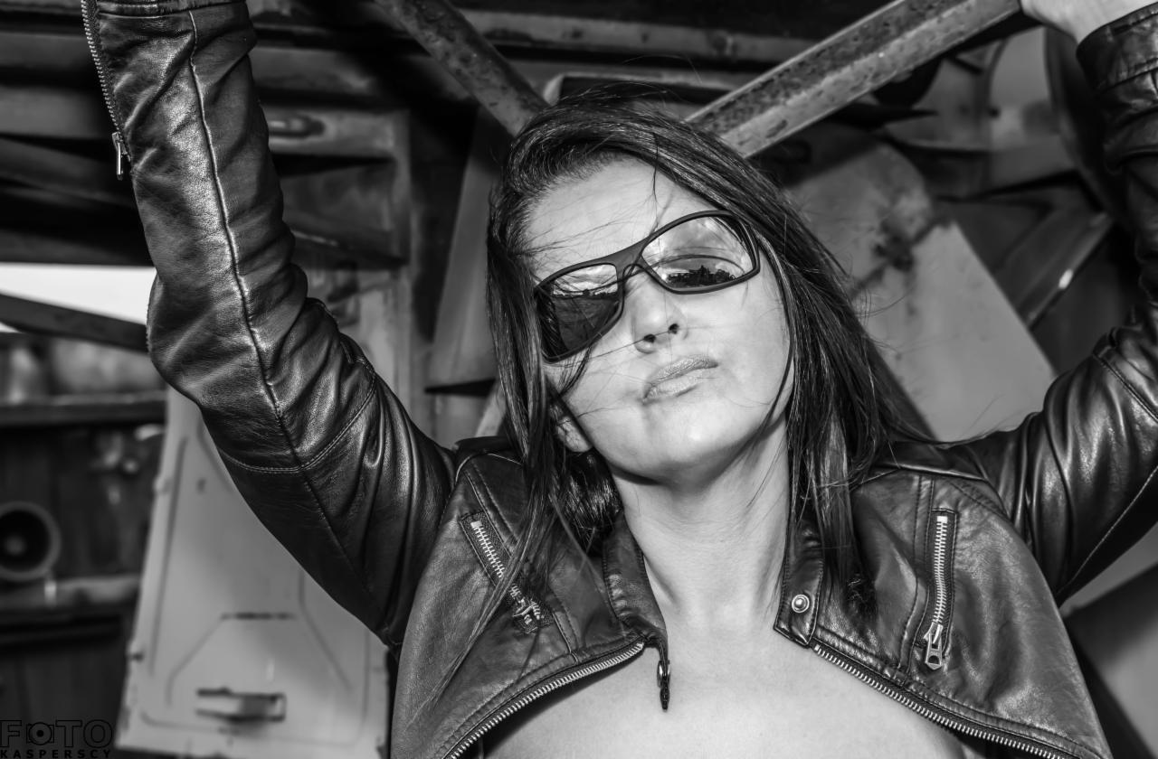 Czarno-białe zdjęcie kobiety w skórzanej kurtce i okularach przeciwsłonecznych, z wiatrem we włosach, pozującej na tle metalowej konstrukcji.