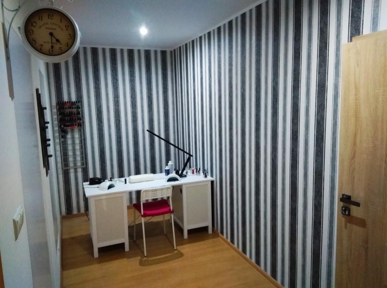 Wnętrze gabinetu kosmetycznego z białym biurkiem do manicure, lampą LED, czerwonym krzesłem na tle ściany w czarno-białe pasy i zegarem w stylu retro.