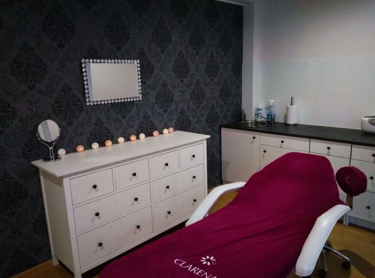 Wnętrze gabinetu kosmetycznego: biała komoda z lustrem w ozdobnej ramie, fotel kosmetyczny przykryty bordowym pokrowcem z logo Clarena, umywalka i szafki na tle ciemnej ściany w ornamenty.