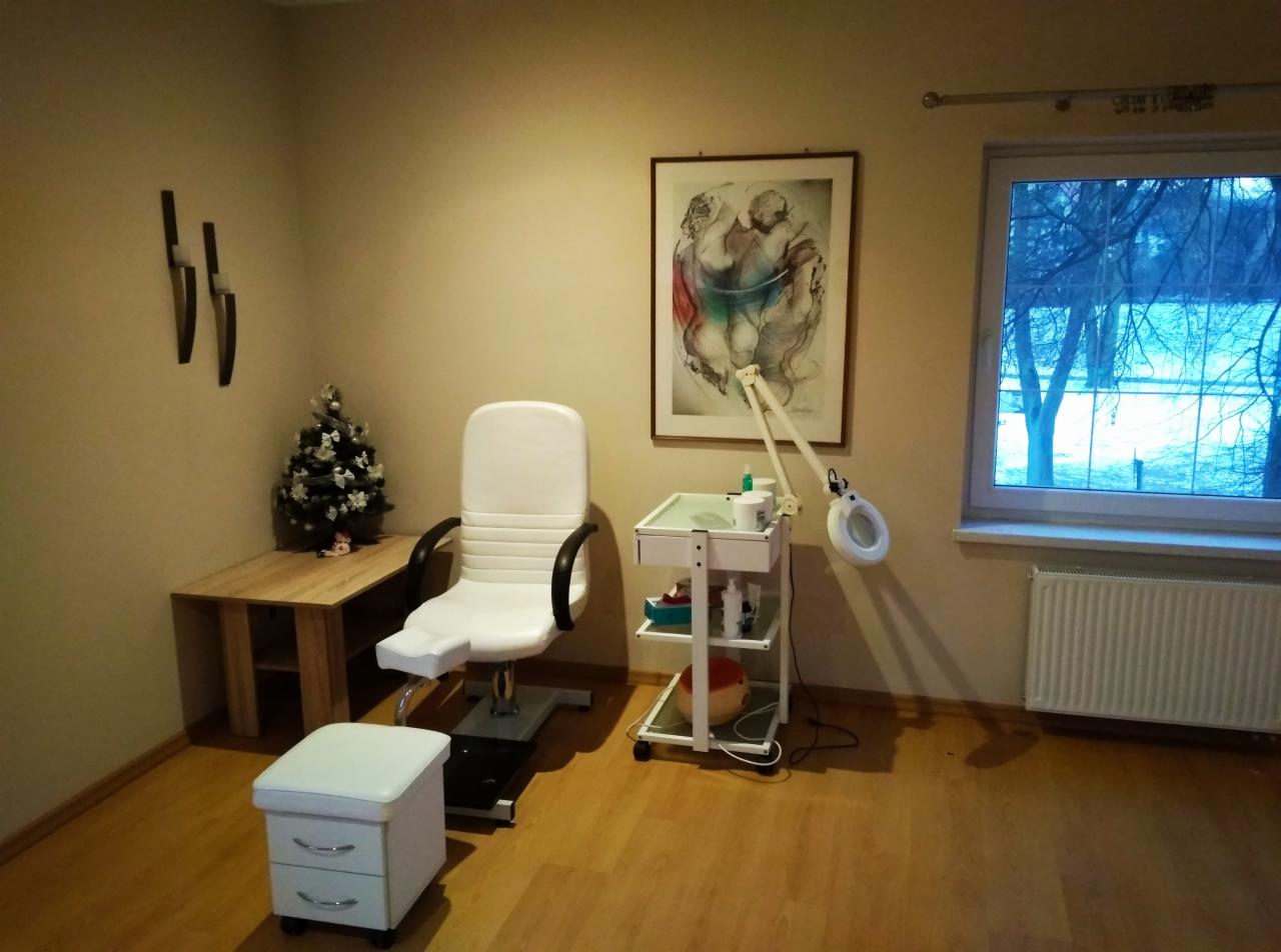 Wnętrze gabinetu kosmetycznego z białym fotelem zabiegowym, stolikiem na kółkach z lampą powiększającą, małą choinką na drewnianym stoliku i obrazem na ścianie.