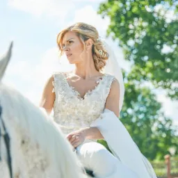 Panna młoda w koronkowej sukni i welonie siedzi na białym koniu, spoglądając w bok na tle zieleni.