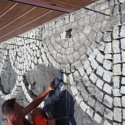 Pracownik układa półokrąg z kostki granitowej na podbudowie z piasku, wyznaczony sznurkami, widok z góry na placu budowy.