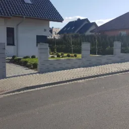 Nowoczesne ogrodzenie z szarej kostki betonowej przed domem z numerem 21 i zadbanym trawnikiem, widok z ulicy.