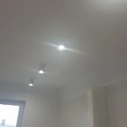 Biały sufit z trzema okrągłymi lampami punktowymi LED, widok z dołu, ściana z płyt kartonowo-gipsowych, okno z widokiem na budynek z cegły.