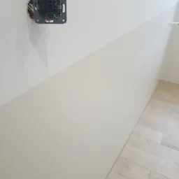 Biała ściana z częściowo wykończonymi panelami ściennymi i wystającą puszką elektryczną bez gniazdka, drewnopodobna podłoga z płytek w prawym dolnym rogu.