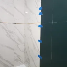 W trakcie układania płytek w łazience: białe płytki imitujące marmur po lewej stronie, ciemnozielone płytki po prawej, pionowa linia styku zabezpieczona niebieską taśmą malarską, widoczna wanna...